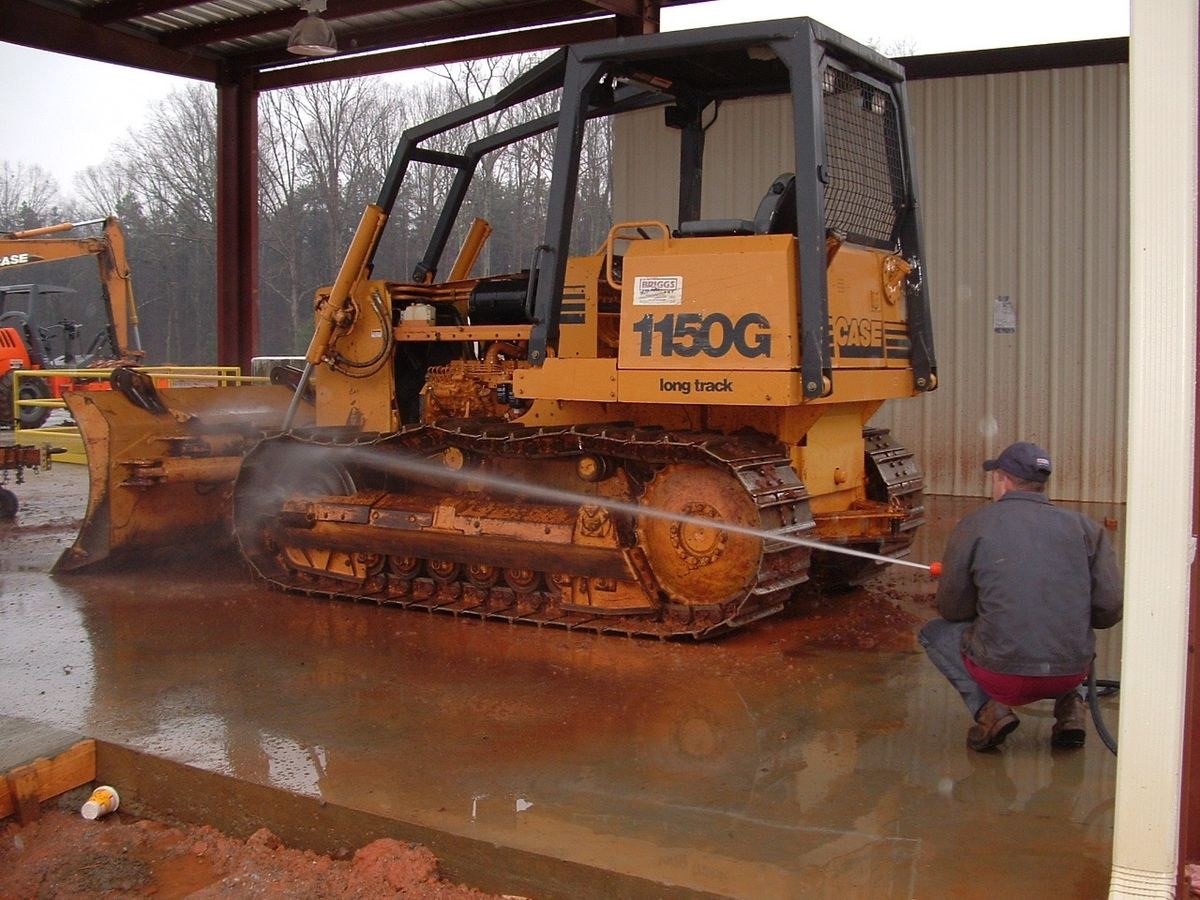 Worker pressure washing mud off a yellow bulldozer at an equipment wash bay